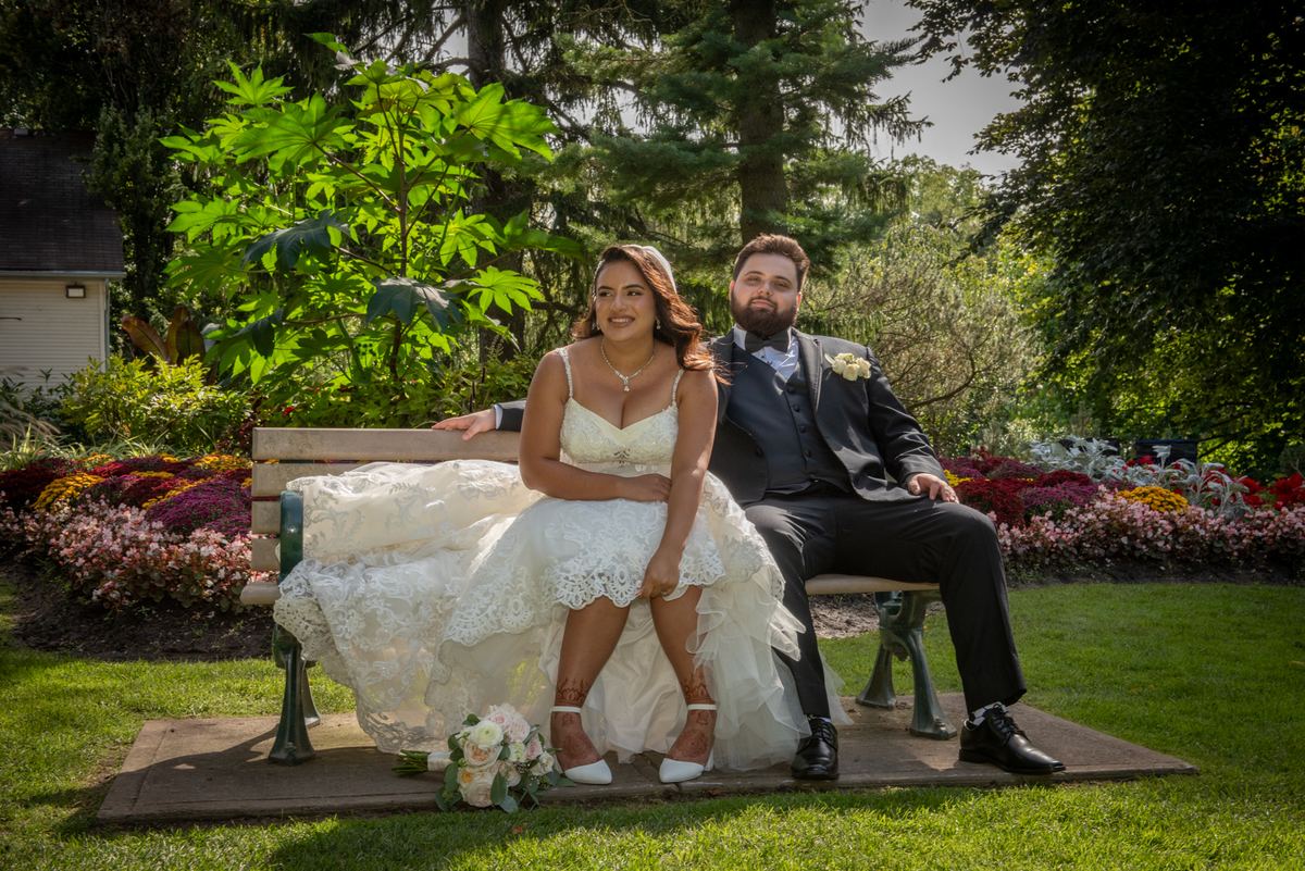 A newlywed couple in formal attire sits on a garden bench, smiling as sunlight filters through the trees.