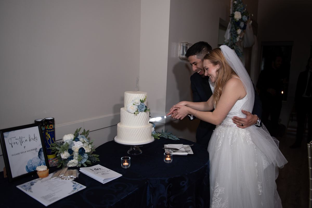 A bride and groom laugh as they cut their multi-tiered wedding cake adorned with blue and white flowers.