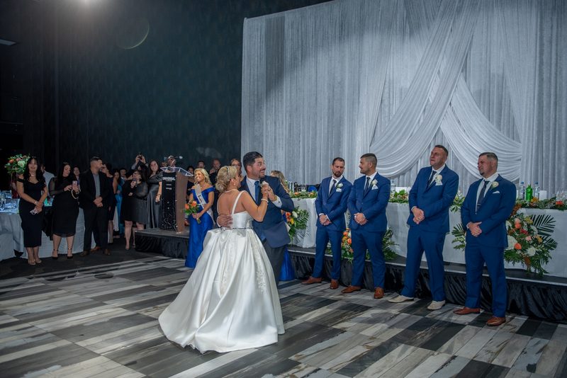 A bride and groom hold each other in their first dance under a spotlight as guests and groomsmen in blue suits applaud.