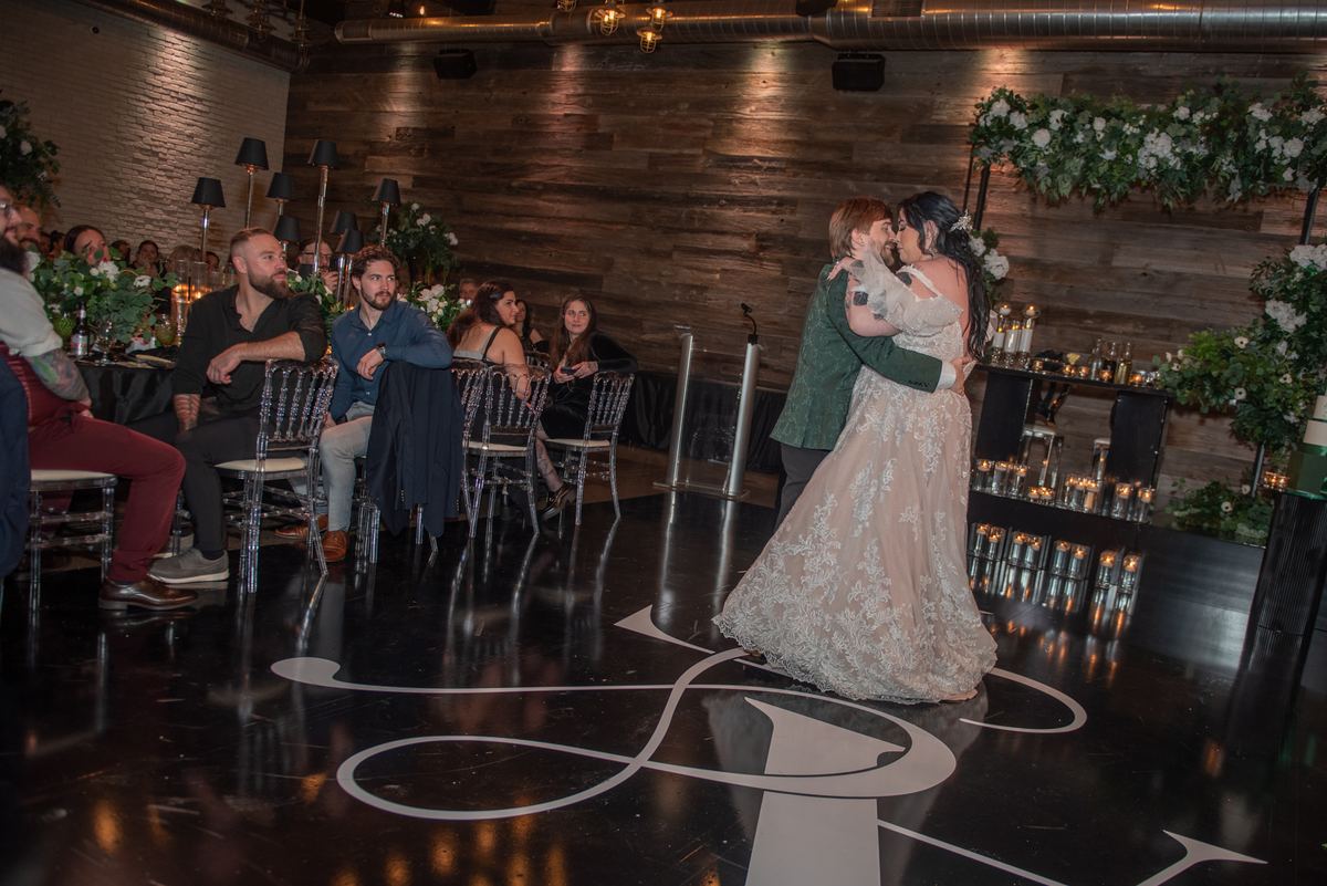 A bride and groom dance on a glossy black floor as guests smile from wooden tables adorned with candles and flowers.
