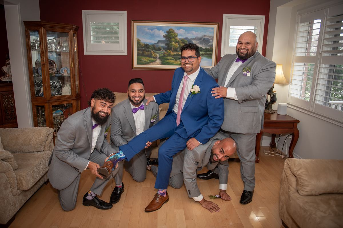A groom is lifted by his groomsmen in a joyful moment, dressed in vibrant suits against red walls and wooden floors.