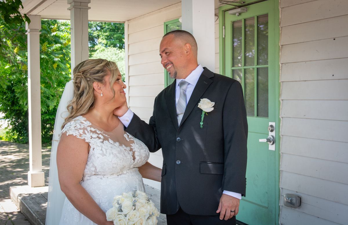 A newlywed couple in bright daylight on a porch, the bride in a lace gown and veil with white roses.