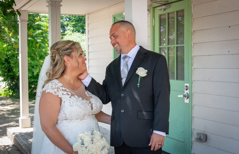 A newlywed couple in bright daylight on a porch, the bride in a lace gown and veil with white roses.