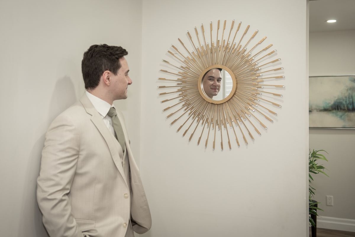 A man in a light beige suit stands gazing at his reflection in a sunburst mirror on a white wall, contemplating.