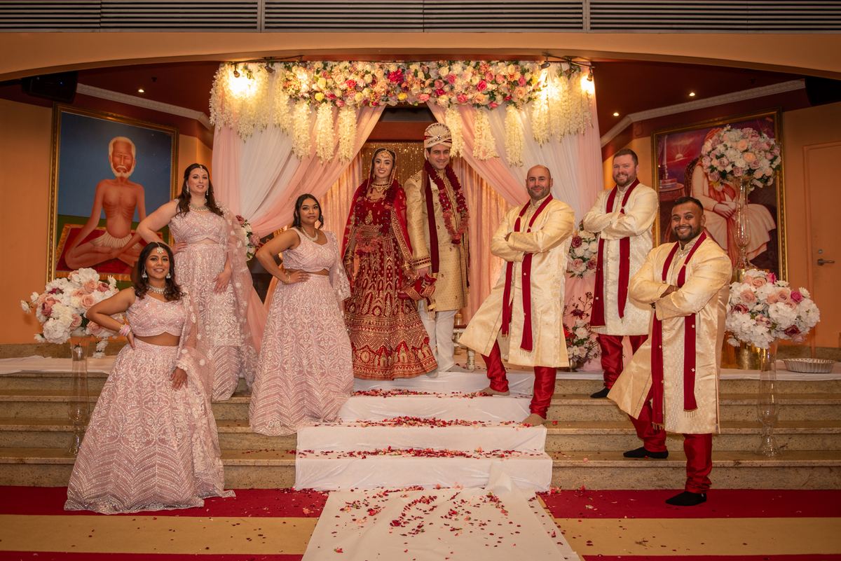 A South Asian wedding group portrait with the bride and groom at center, surrounded by bridesmaids in pink lehengas.