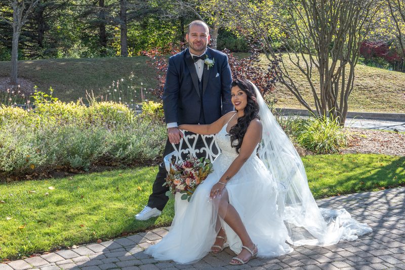 A bride in a white dress and groom in a suit stand beside each other near a decorative bench under trees on a sunny day.