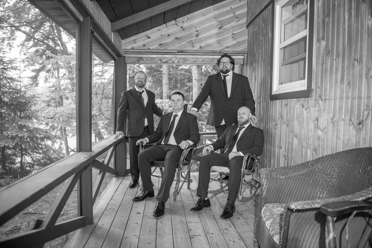 Four men in suits pose on a wooden porch, three seated and one standing, with soft natural light through trees in background.