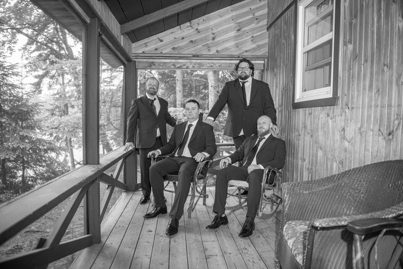 Four men in suits pose on a wooden porch, three seated and one standing, with soft natural light through trees in background.