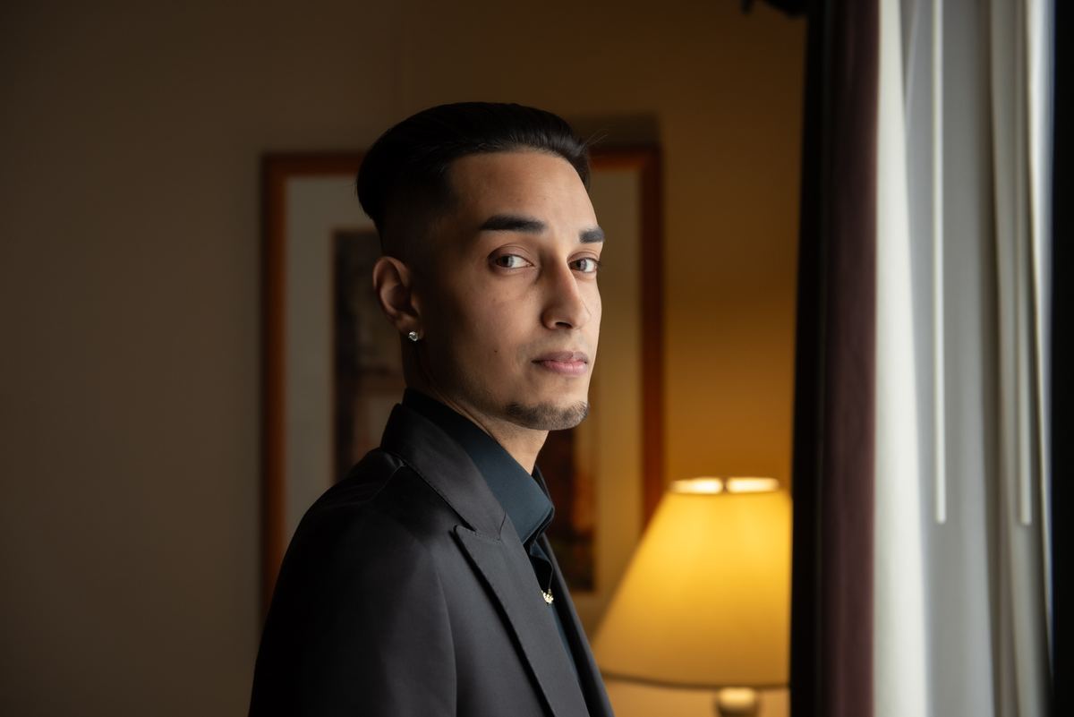 A man in a dark suit stands near a window, gazing thoughtfully, with warm indoor light contrasting cool natural light.