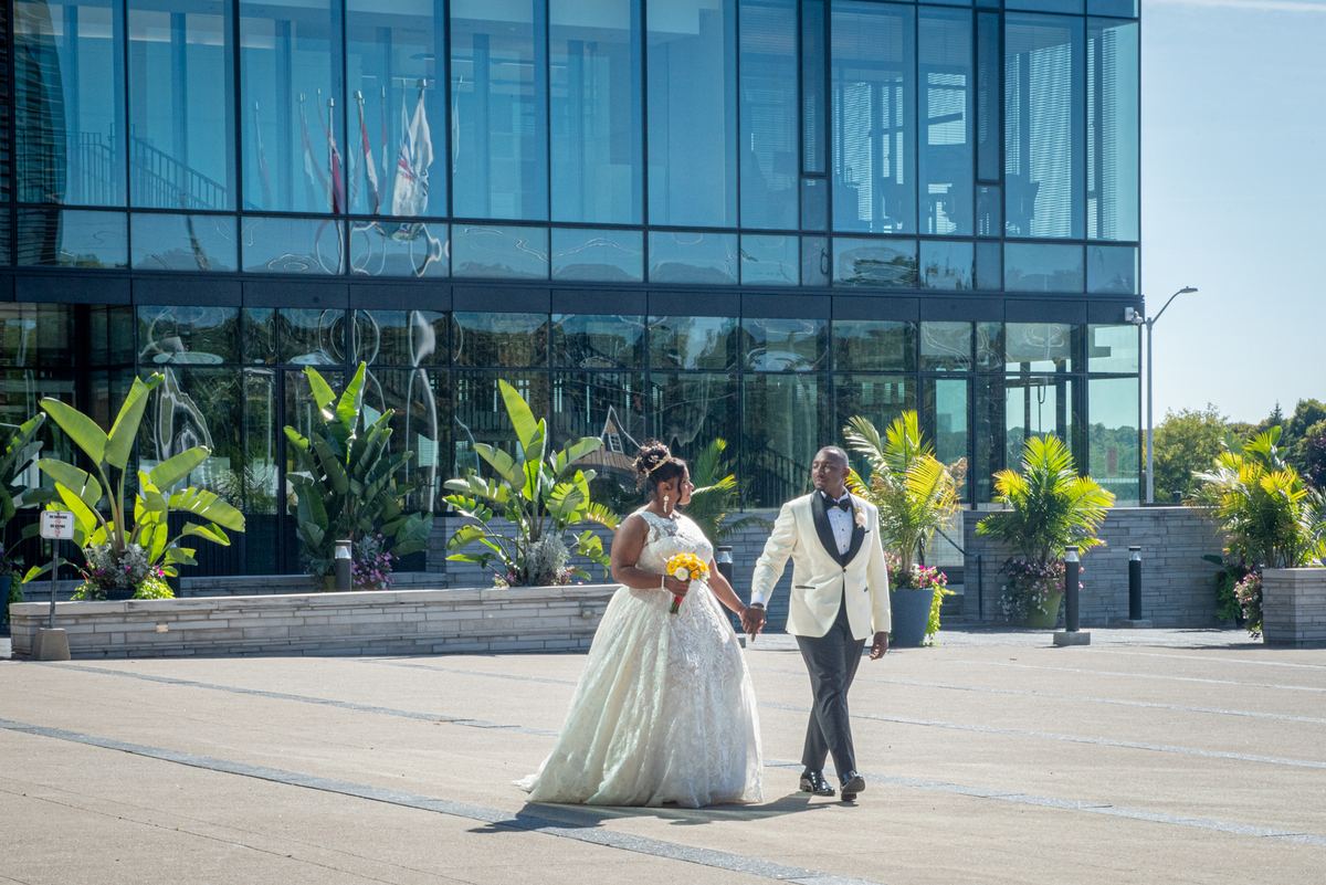Gabrielle and Elkanah walk hand-in-hand across a paved plaza in front of a modern glass building, bathed in bright sunlight.