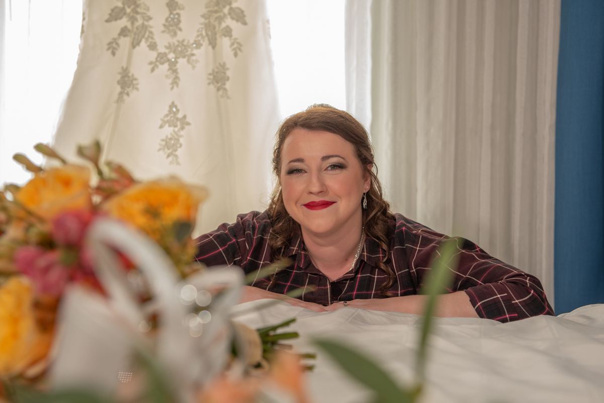 A bride in casual attire smiles softly at a table with flowers and sheer curtains behind her, filtering soft light.