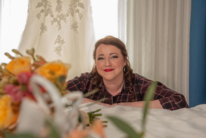 A bride in casual attire smiles softly at a table with flowers and sheer curtains behind her, filtering soft light.