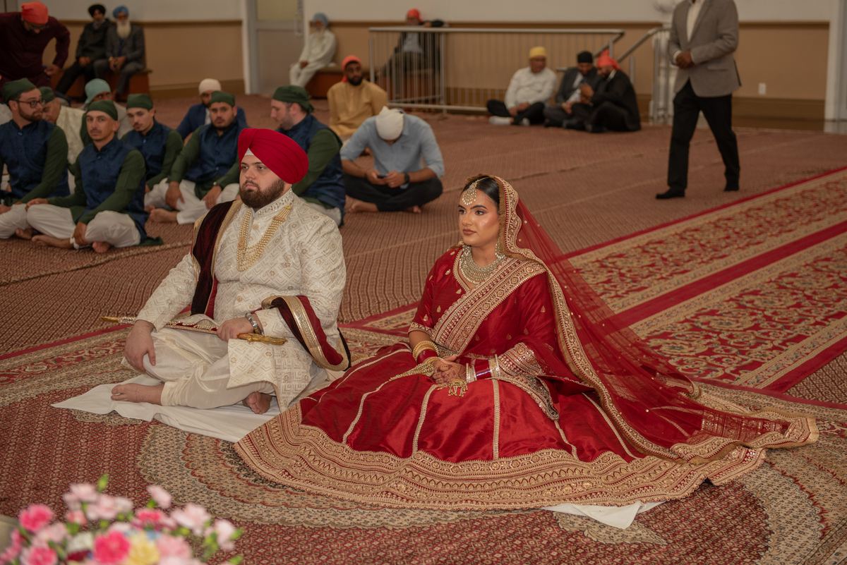 A Sikh bride and groom sit on an ornate rug during a wedding ceremony, surrounded by guests in traditional attire.