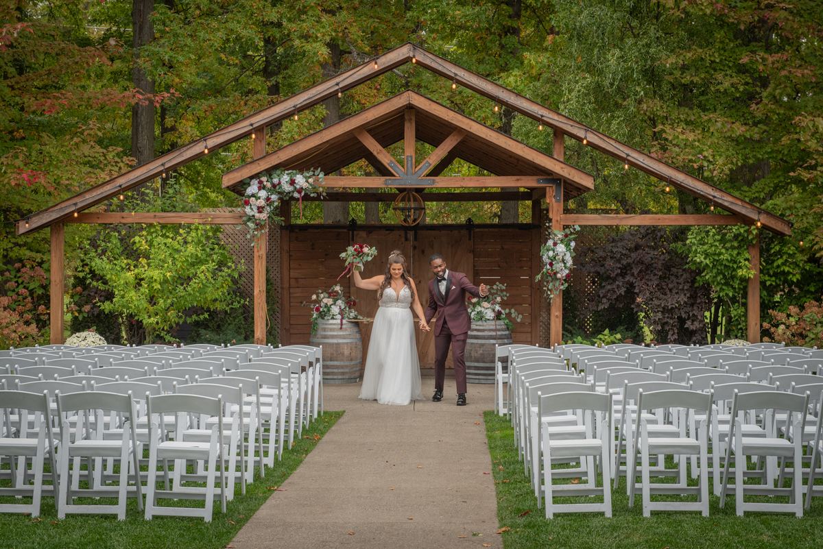 A smiling bride and groom walk hand-in-hand down the aisle under a rustic wooden archway adorned with flowers.