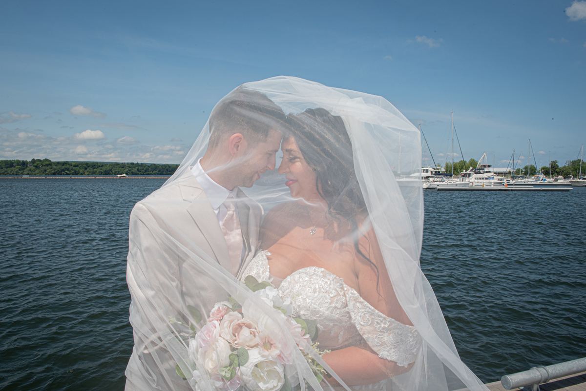 Bride and groom share tender moment on waterfront dock, foreheads touching, under partly cloudy sky.