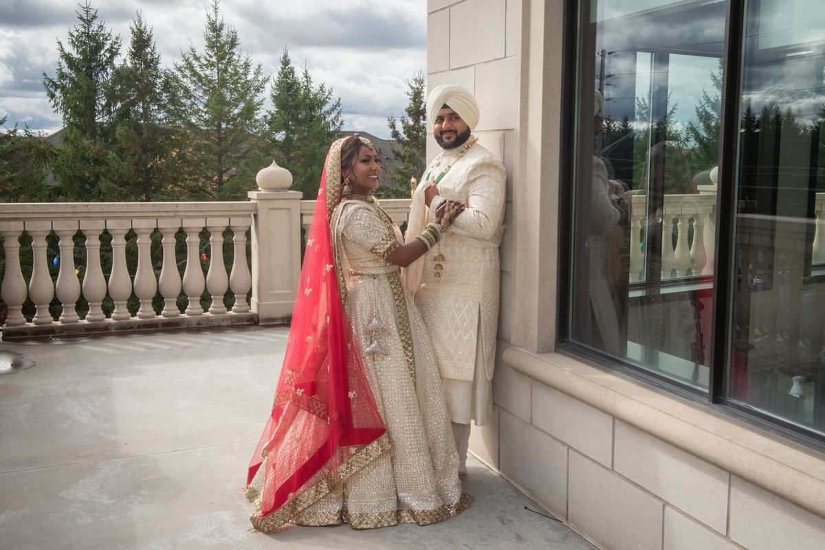 A bride and groom in traditional South Asian attire stand on a stone balcony, hands clasped, smiling.