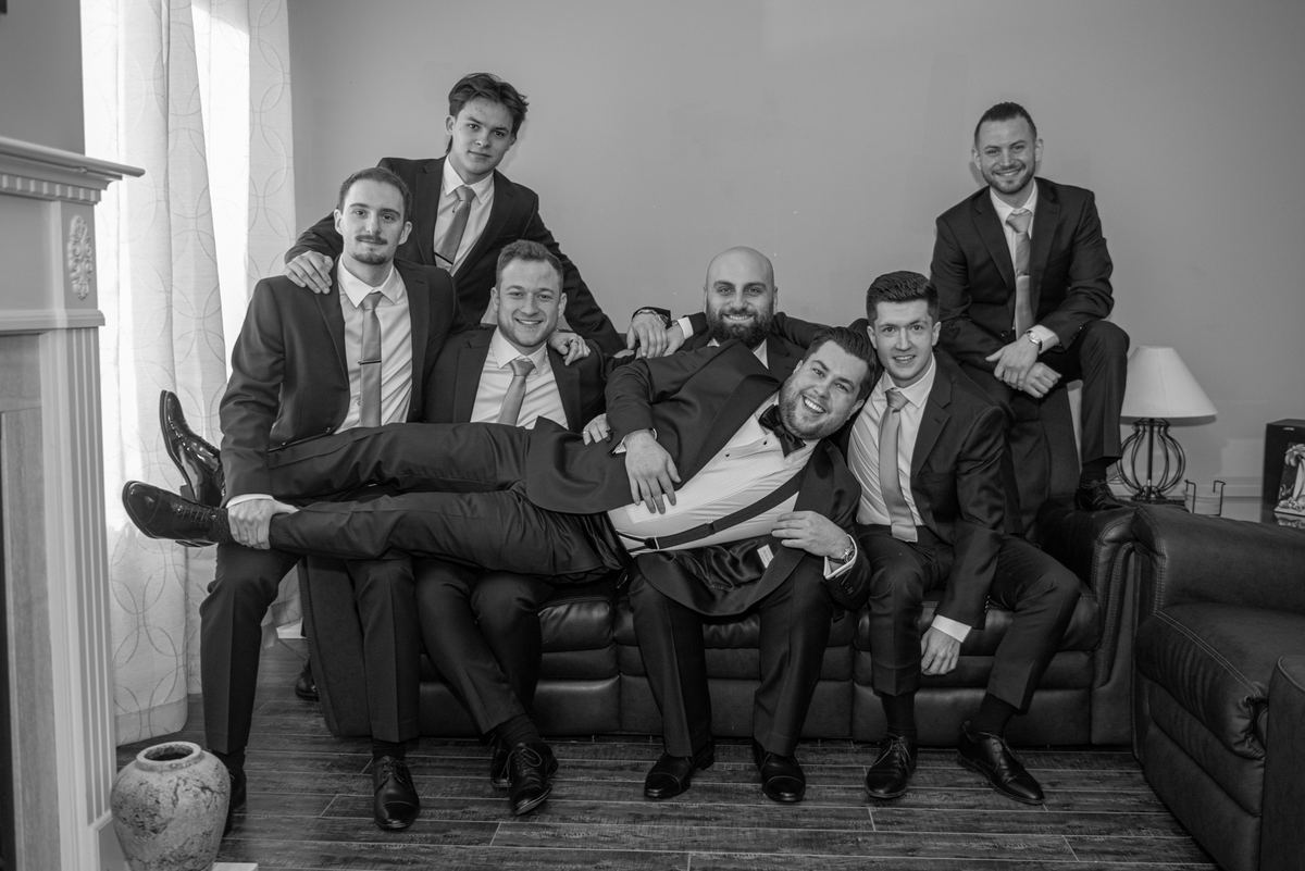 Seven formally dressed men pose playfully on a leather sofa with natural light streaming in through a window.