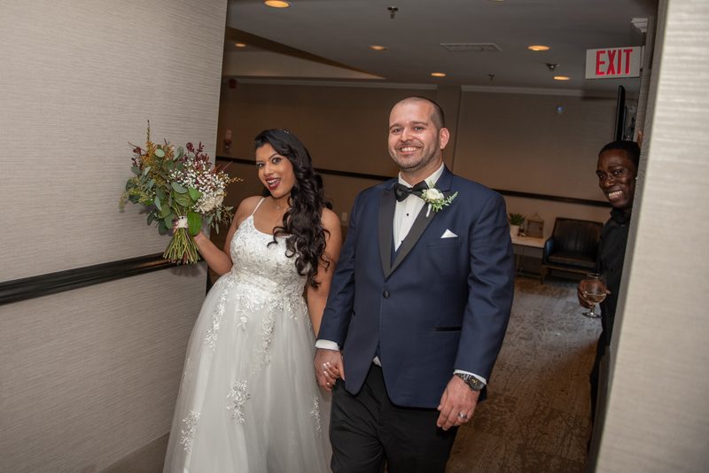 A smiling bride and groom hold hands, walking down a corridor with warm lighting and an EXIT sign overhead.
