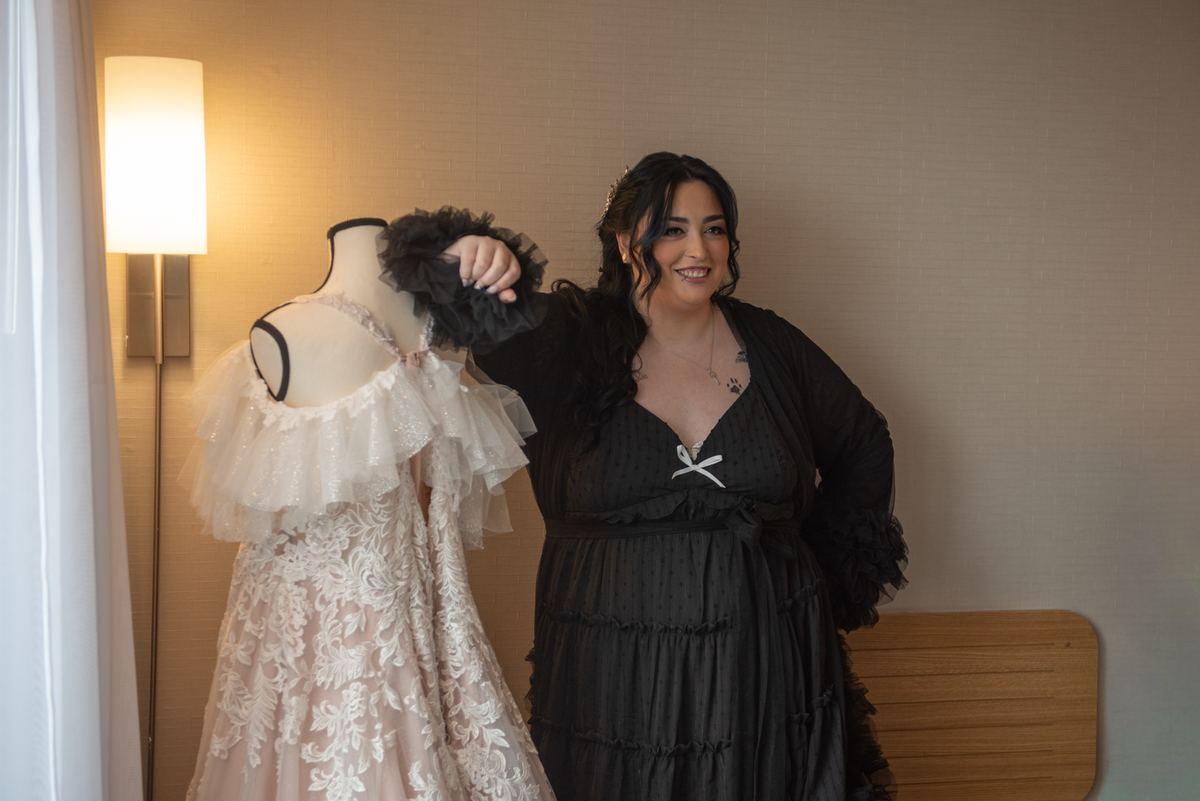 A woman smiles beside a mannequin in an ornate lace gown, illuminated by warm light from a wall sconce.