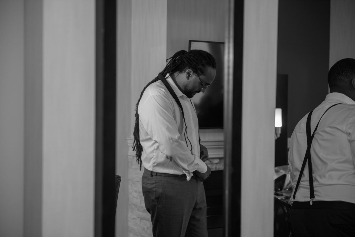 Two formally dressed men prepare in a dimly lit room, one adjusting his clothing while the other waits.