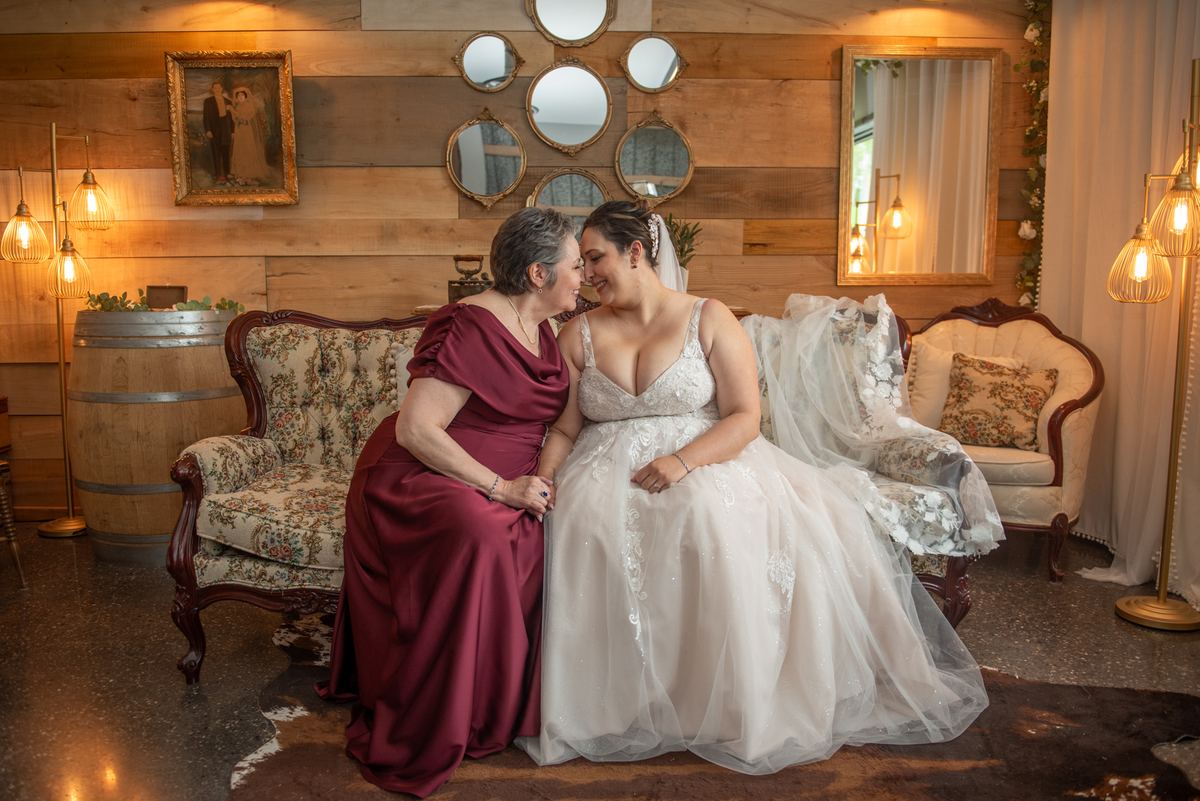 A bride in a white gown sits closely with a family member in burgundy on ornate floral sofas, smiling.
