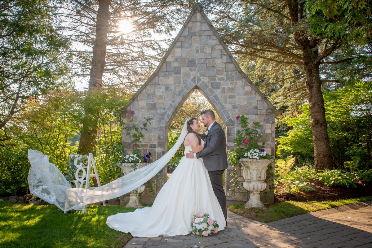 A couple stands in a Gothic archway, framed by trees and planters, as golden light highlights their intimate moment.