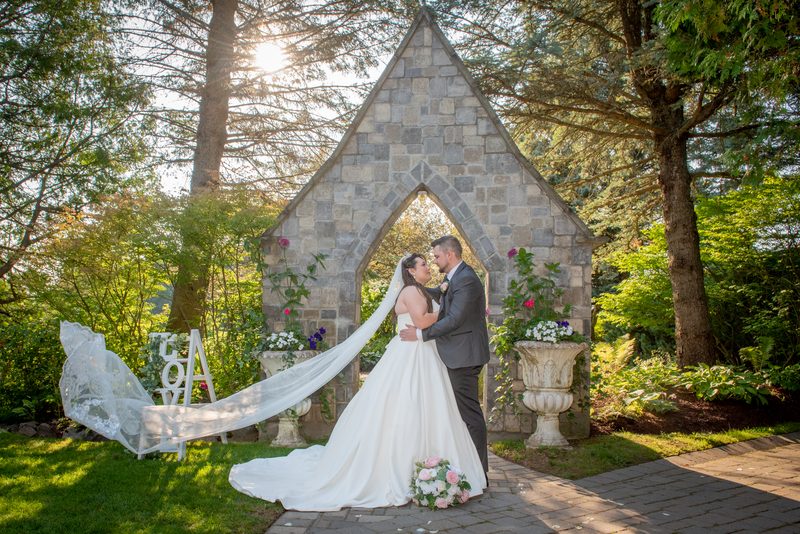 A couple stands in a Gothic archway, framed by trees and planters, as golden light highlights their intimate moment.