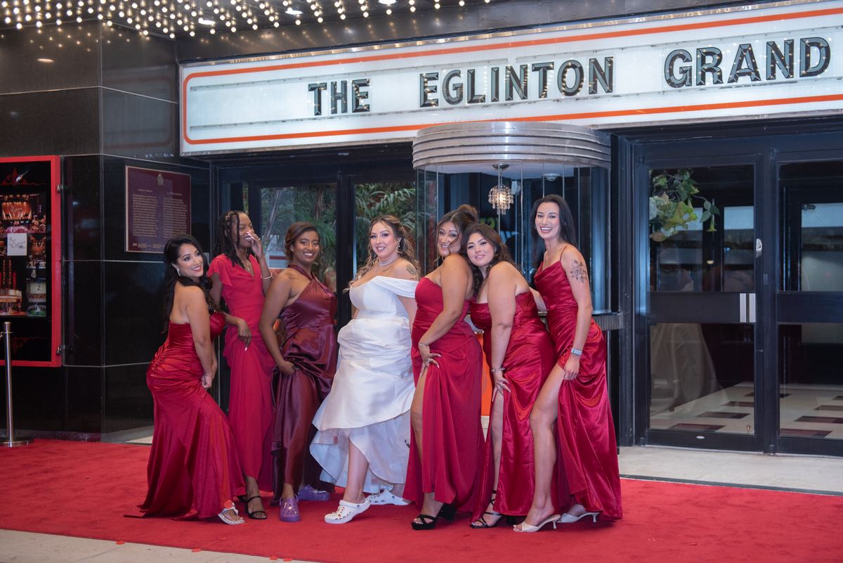 A bride in white stands with her red-clad bridesmaids on a red carpet, illuminated by fairy lights and warm lighting.
