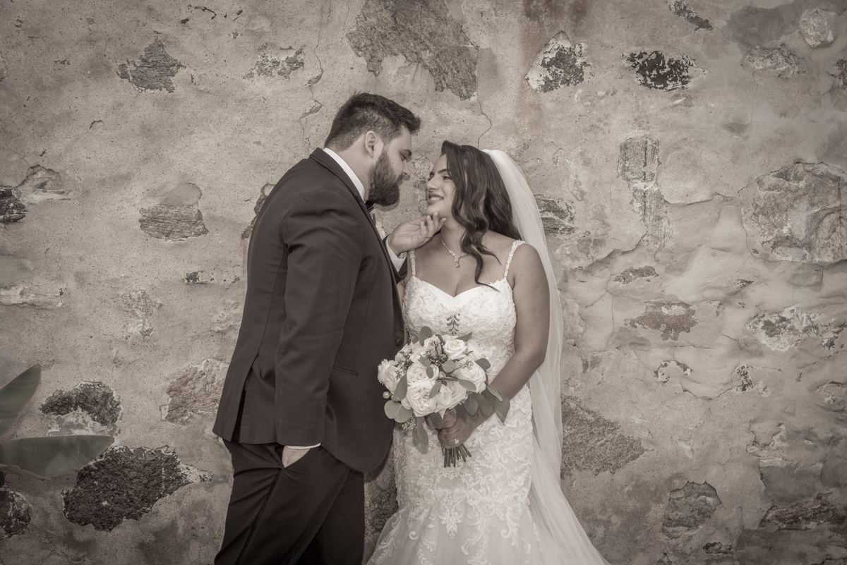 A bride and groom share a tender moment by a weathered stone wall, the groom touching her cheek as she holds a bouquet.