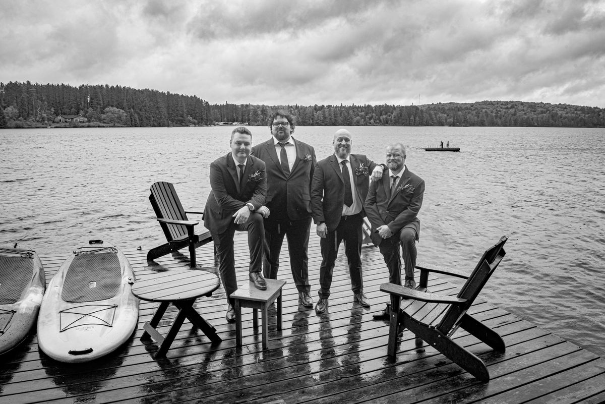 Four men in suits stand on a wooden dock by a lake, with paddleboards nearby and a distant shoreline visible.
