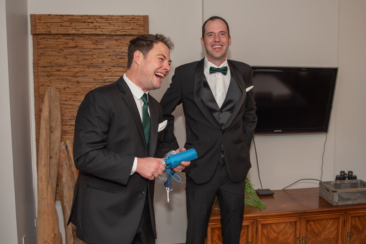 Two men in tuxedos with green bow ties laugh heartily, one holding a small blue umbrella, under bright indoor lighting.