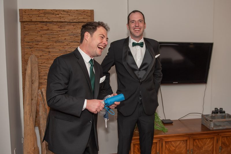 Two men in tuxedos with green bow ties laugh heartily, one holding a small blue umbrella, under bright indoor lighting.
