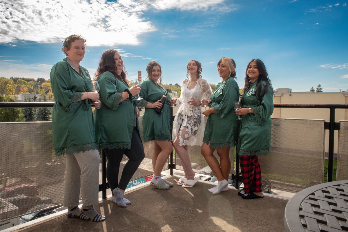 A bride and her bridesmaids laugh and toast on a sunny rooftop terrace with a view of trees and buildings.