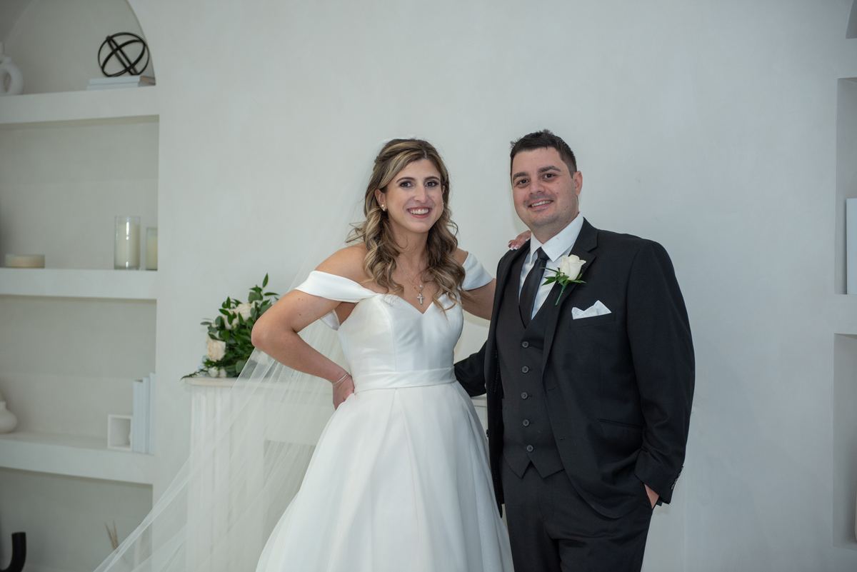 A smiling bride and groom in formal attire pose joyfully in a minimalist white room during a wedding photoshoot.