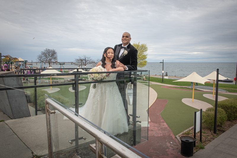 A newlywed couple smiles at the camera from an overlook with glass railings, overlooking a waterfront park.