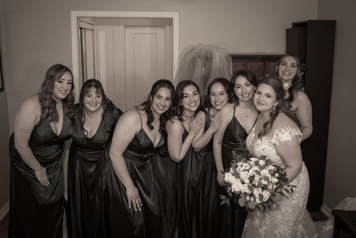 A bride in a lace gown with her bridesmaids in matching satin gowns smiles indoors near a draped veil.
