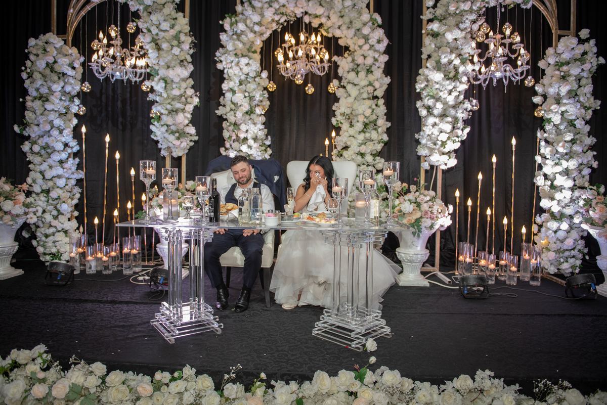 A bride and groom sit at a clear table under cascading floral arches, illuminated by chandeliers and candles.