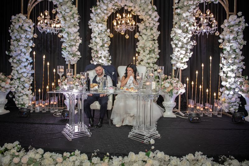 A bride and groom sit at a clear table under cascading floral arches, illuminated by chandeliers and candles.