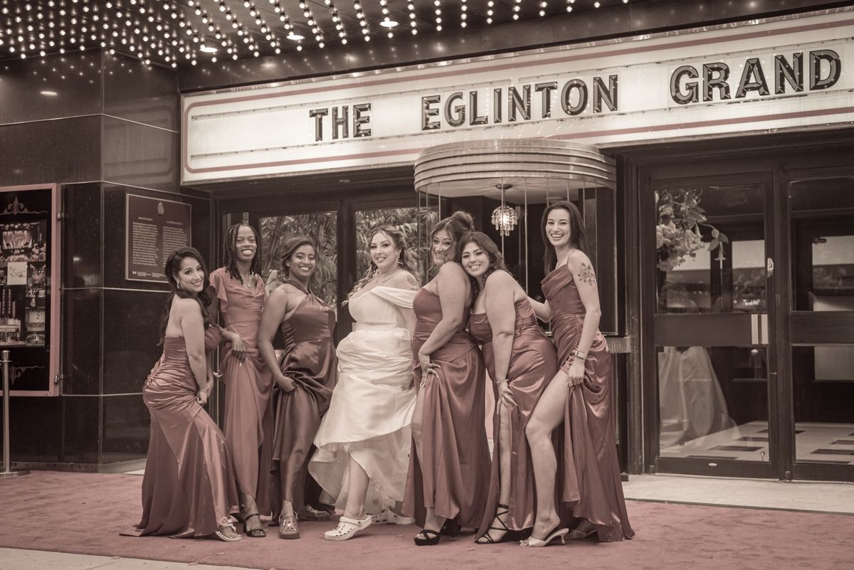 A bride and her bridesmaids pose joyfully in front of a historic theater, illuminated by warm string lights and the marquee.