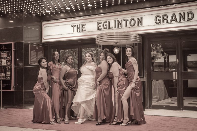 A bride and her bridesmaids pose joyfully in front of a historic theater, illuminated by warm string lights and the marquee.