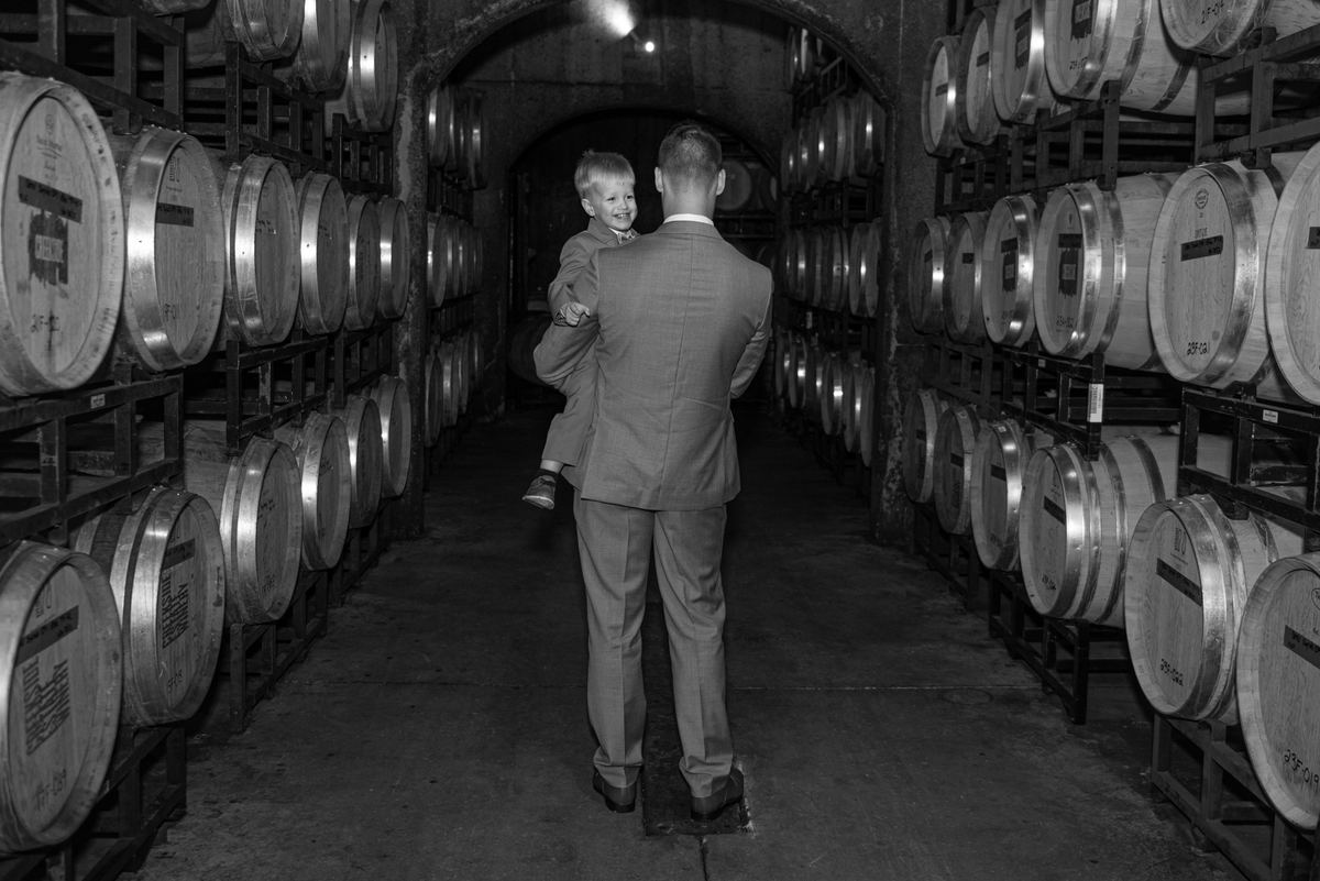 A man in a suit carries a child through a dim cellar with wooden barrels and stone walls, illuminated by soft light.