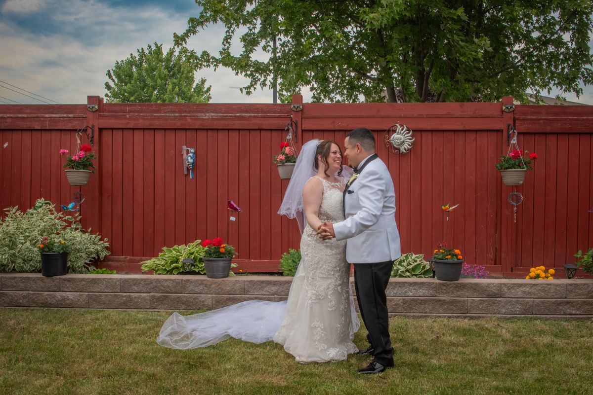 Bride and groom stand on grass, holding hands and gazing at each other, with a red fence decorated with flowers behind them.