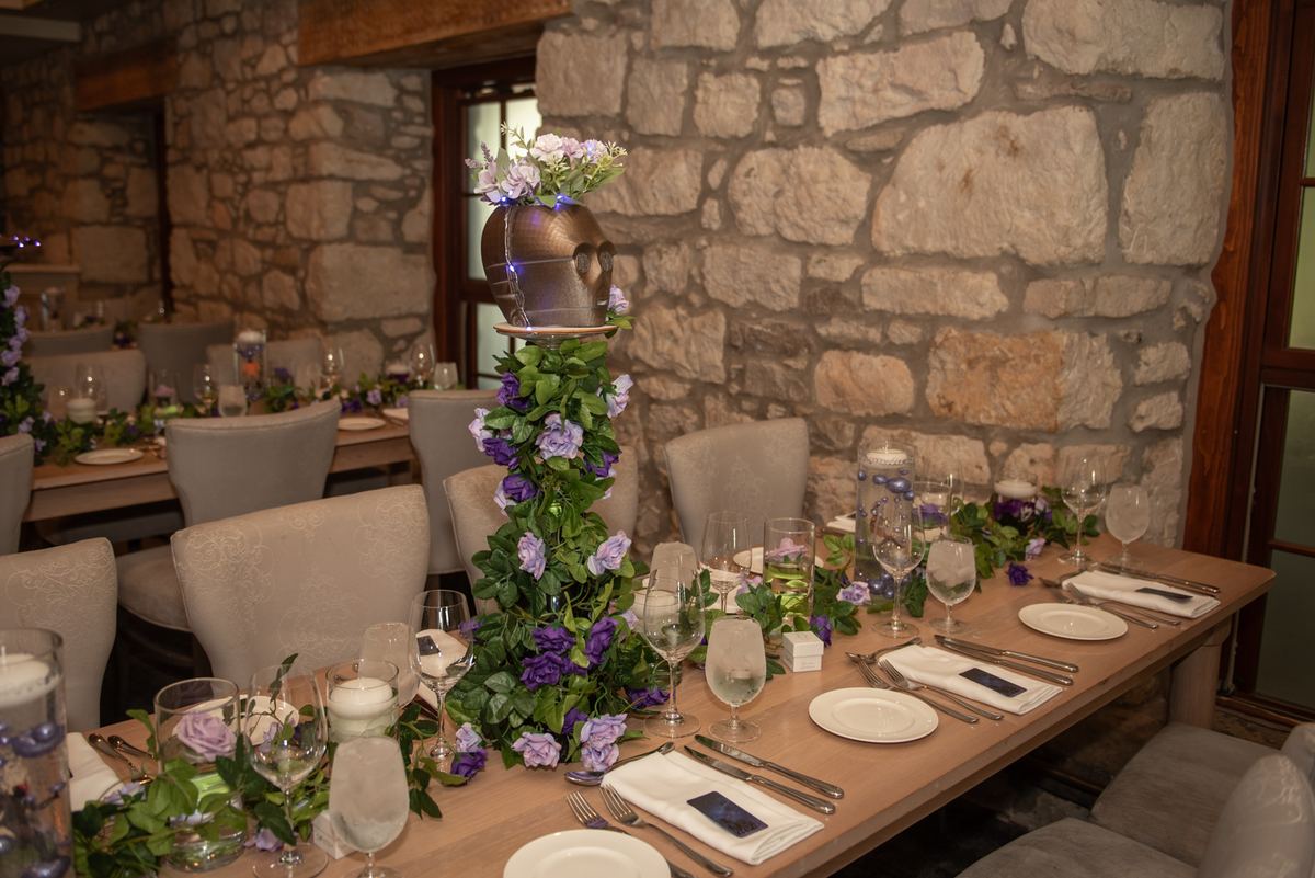 A rustic dining table set with floral arrangements and candles under soft light in a stone-walled room.
