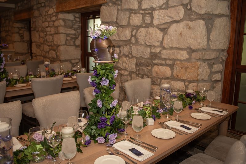 A rustic dining table set with floral arrangements and candles under soft light in a stone-walled room.