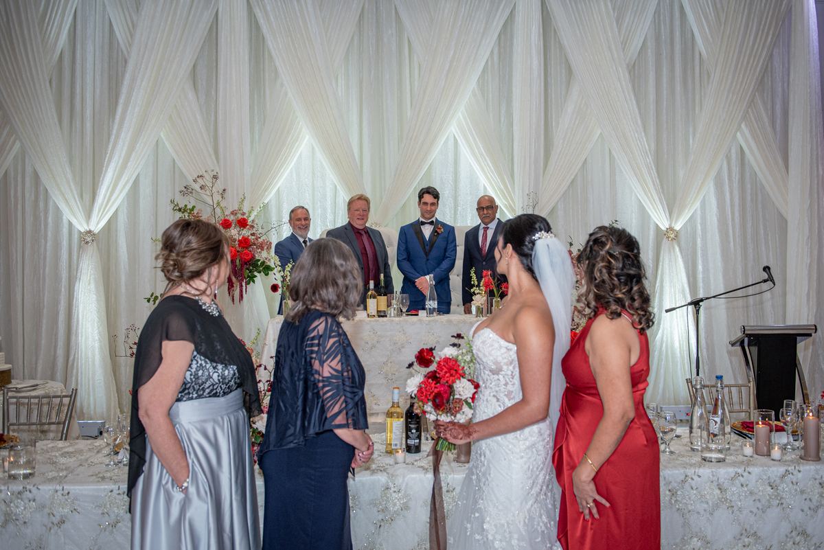 A bride in a white dress holds flowers as she faces a table with family members and guests in formal attire.