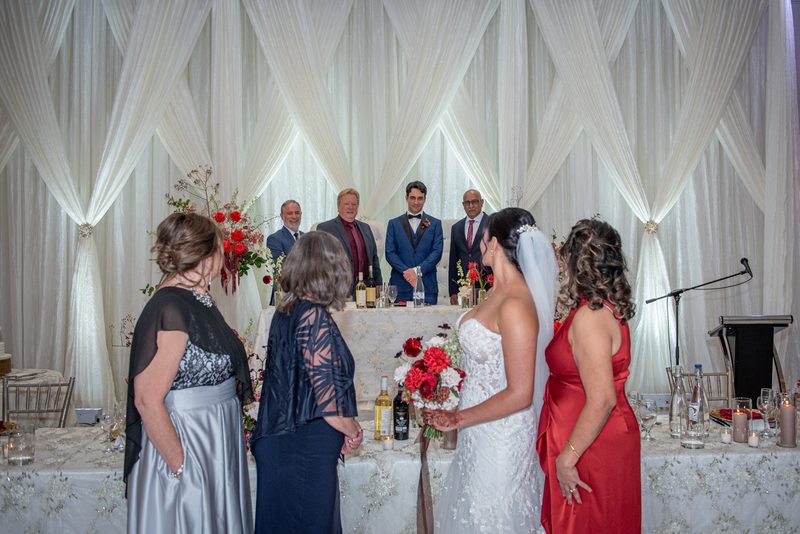 A bride in a white dress holds flowers as she faces a table with family members and guests in formal attire.
