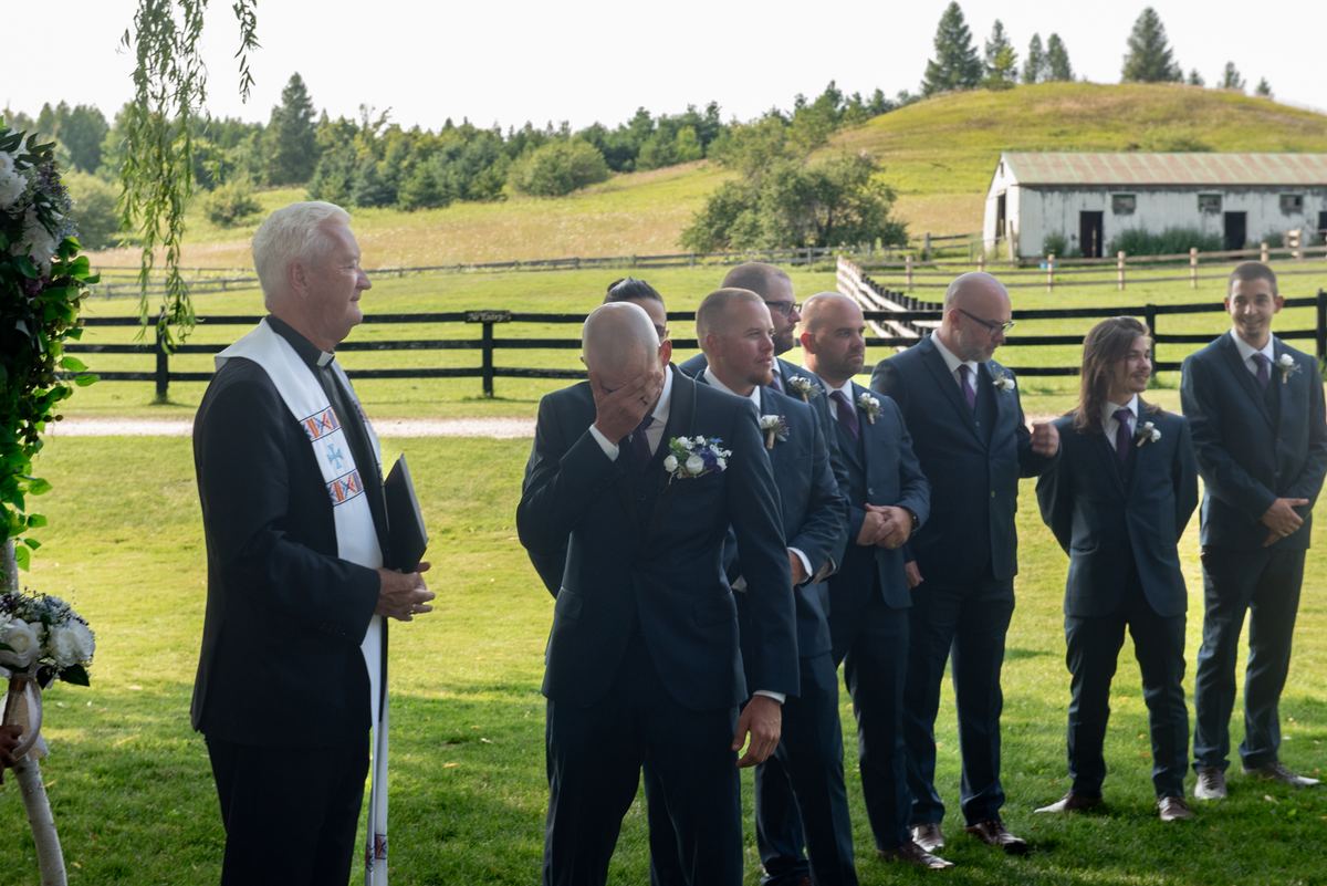 An emotional groom covers his face during an outdoor wedding ceremony near a wooden fence and rolling hills.