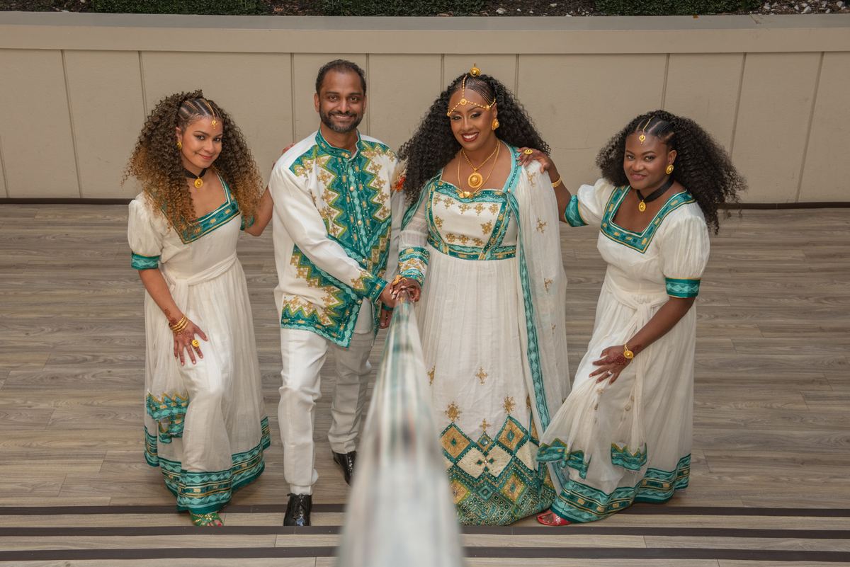 Four people in traditional African attire smile warmly in a tilted photograph with blurred fabric in foreground.