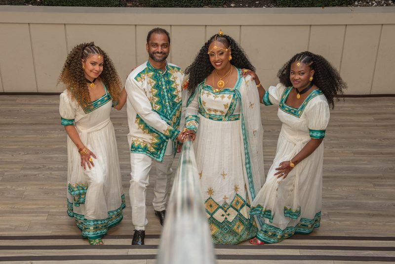 Four people in traditional African attire smile warmly in a tilted photograph with blurred fabric in foreground.