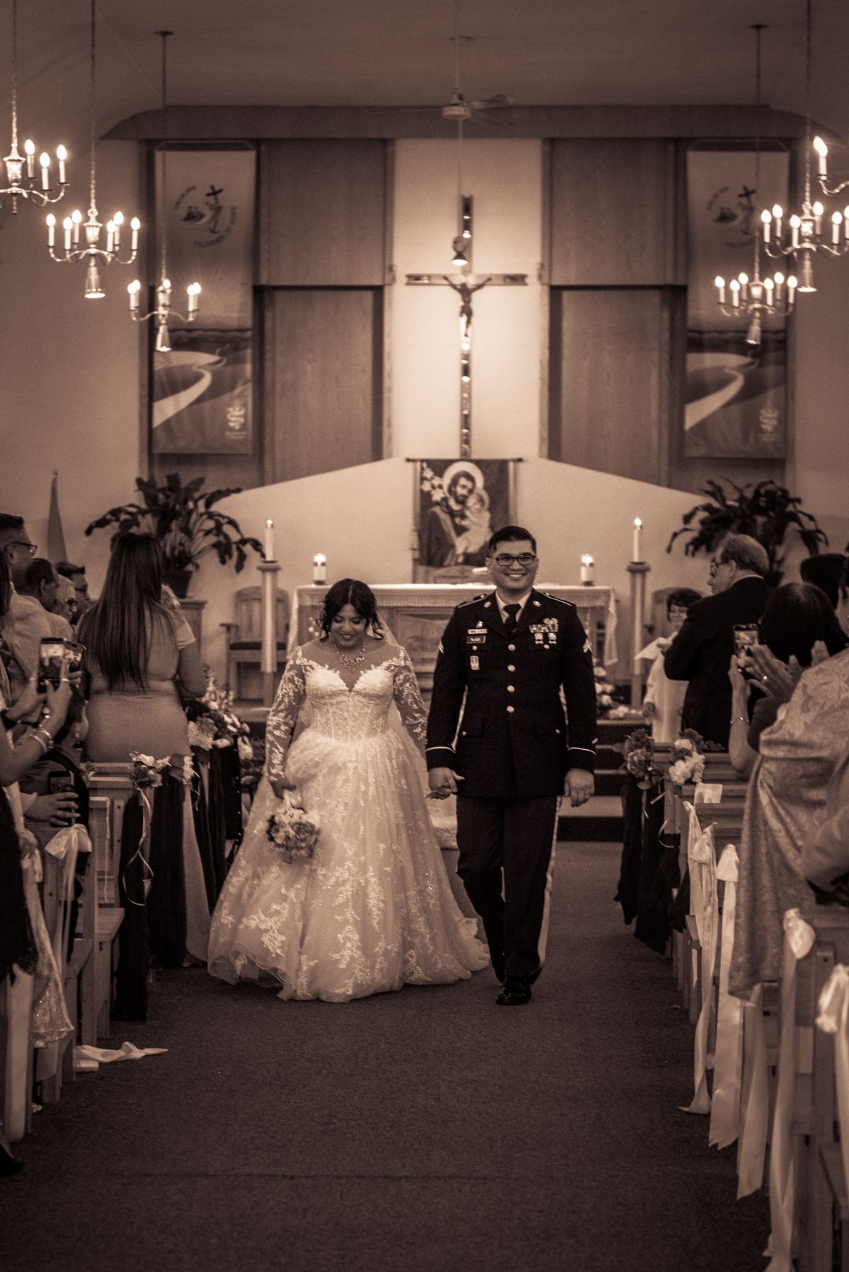 A bride in a lace gown and groom in military uniform walk down the church aisle, surrounded by guests.